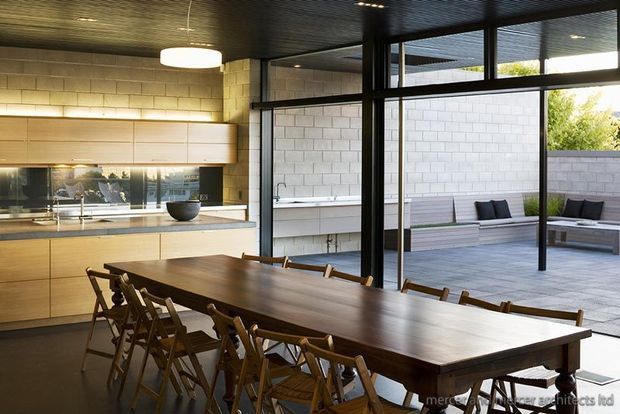 A warm, contemporary open-plan kitchen and dining area with extensive black-framed glazing opening onto a paved courtyard. The kitchen features light timber cabinetry, a dark stone or composite benchtop, integrated lighting, and a long solid timber dining table with folding timber chairs. The courtyard includes built-in pale seating against a light masonry wall. The interior has a dark ribbed ceiling and strong indoor-outdoor connection.