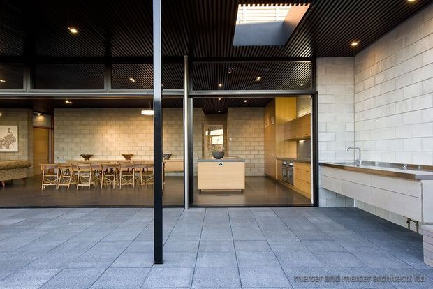 A spacious contemporary kitchen and dining area viewed through large black-framed glazed openings. The kitchen features light timber cabinetry, a central island, stainless-steel work surfaces and sink, pale masonry block walls, and a dark ribbed ceiling with recessed lighting and a skylight. A long dining table and chairs sit to the left, while large grey floor tiles extend across the foreground.