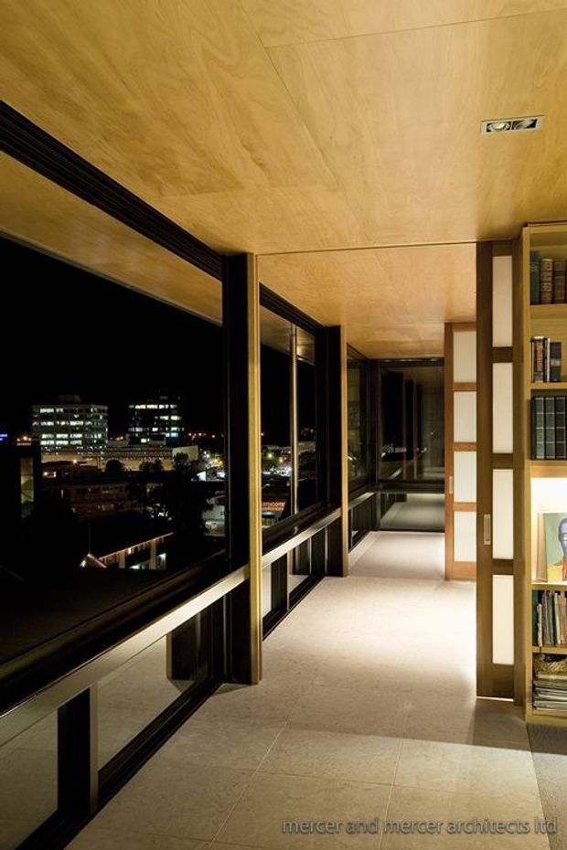 A nighttime view along a refined interior corridor or landing with expansive black-framed floor-to-ceiling glazing overlooking a city. The space features a warm timber-lined ceiling, pale stone or tile flooring, dark window frames, integrated recessed lighting, and a built-in timber bookcase with illuminated shelving. Reflections in the glazing extend the corridor visually, creating a sleek, calm architectural interior.