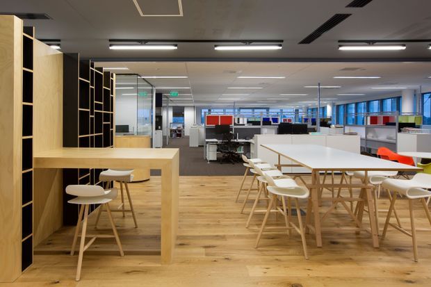 Open-plan contemporary office interior with timber flooring, pale timber communal tables and stools, white workstations, black-framed timber shelving, glass partitions, and rows of linear ceiling lights. The space includes collaborative breakout seating in the foreground and desk-based work areas beyond, with blue-framed glazing along the rear.
