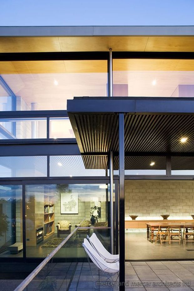 Architectural photograph of a contemporary multi-level living and entertaining space with extensive glazing, dark steel framing, warm timber soffits, and a covered outdoor terrace. A lounge area is visible through the glass on the left, while the right side contains a dining/bar area beneath a slatted ceiling with a long illuminated bench. The image also shows a tiled terrace and white outdoor lounge chairs in the foreground.