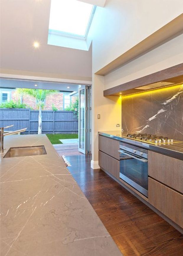 Bright contemporary kitchen with a long stone island and integrated sink, dark wood flooring, dark wood cabinetry, a large veined stone benchtop and splashback, stainless-steel cooking appliances, warm under-cabinet lighting, and a skylight. Bi-fold or fully opening glazing connects the kitchen to a small landscaped courtyard.
