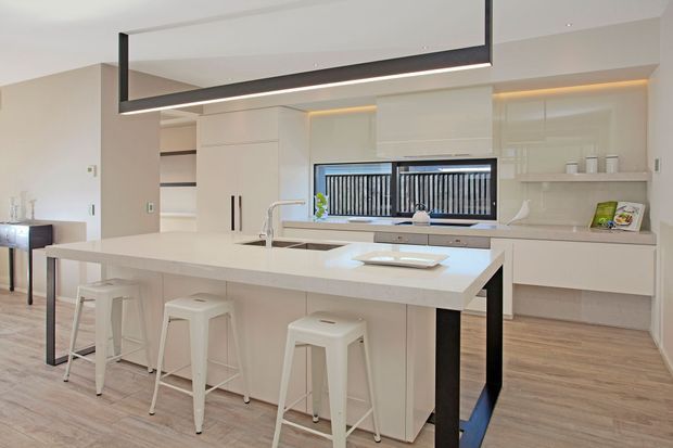 Bright contemporary kitchen with a large white island and integrated sink, four white bar stools, extensive white built-in cabinetry, open shelving, a pale benchtop, timber-look flooring, and a black rectangular pendant light. Horizontal windows with black security screens provide natural light, while warm concealed lighting highlights the cabinetry.
