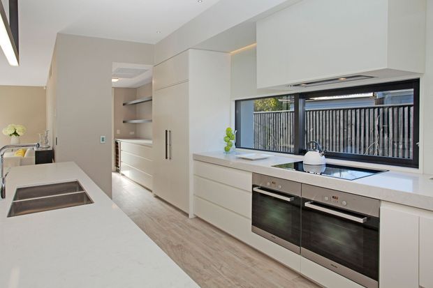 Bright contemporary kitchen with white flat-panel cabinetry, a long white island with integrated dark cooktop, double built-in ovens, pale timber-look flooring, and a wide horizontal window overlooking a slatted privacy fence. A walk-in pantry or storage area is visible beyond the kitchen.