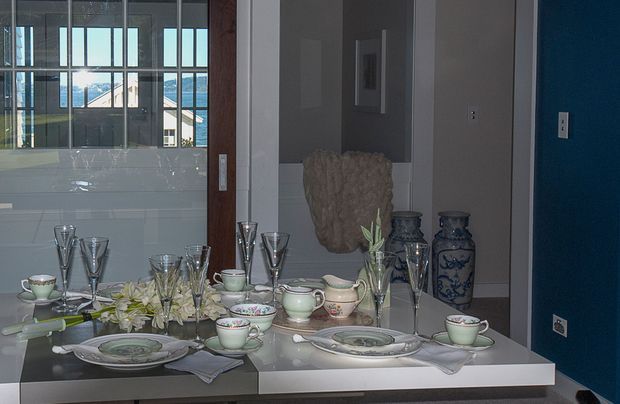 A formal dining setting viewed through a large wall mirror, with a glossy dining table arranged with plates, cups, glassware, floral centrepieces and decorative blue-and-white vases. The room has grey walls, a vivid blue accent wall, and a glazed opening or adjoining space visible in the reflection. The styling is refined and traditional-leaning within a contemporary interior.