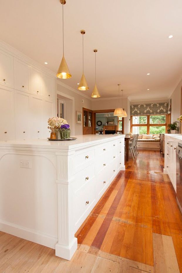 A warm, classic-style kitchen featuring a large white central island with raised-panel cabinetry and brass knobs, extensive matching built-in cabinetry, polished light stone benchtops, timber flooring with contrasting inlaid boards, and a row of gold pendant lights. The kitchen opens toward a dining and living area with timber-framed windows, a window seat, and patterned Roman blinds.