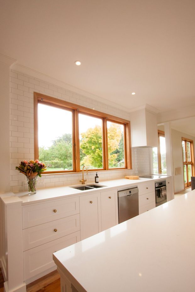 Bright, warm kitchen with white Shaker-style cabinetry, a large white island, white subway-tile walls, timber-framed windows, and stainless-steel appliances. The room features a double-bowl sink beneath a wide bank of windows, recessed ceiling lights, timber flooring, and views of leafy trees outside.