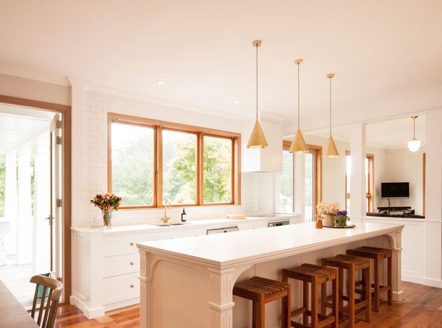 Bright, spacious white kitchen with a large island and seating for four. The room features white brick-look wall tiling, white cabinetry, warm timber-framed windows and doors, pendant lights, timber flooring, and traditional decorative detailing on the island. A living area with a wall-mounted television is visible beyond the kitchen.
