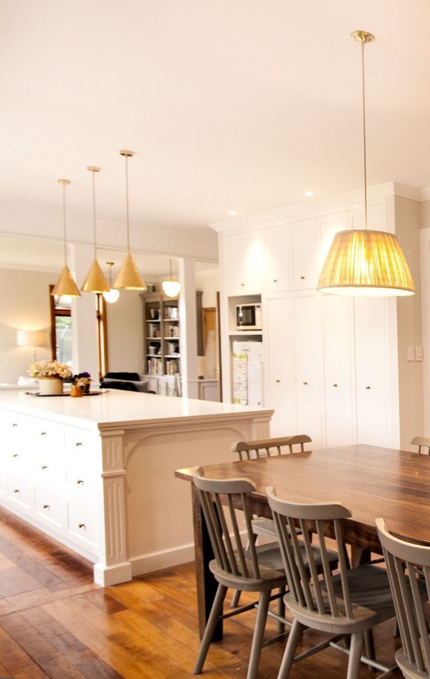 Bright, warm-toned open-plan kitchen and dining area. The kitchen features extensive white built-in cabinetry, a large white island with decorative traditional-style trim, pendant lighting, and integrated appliances. A substantial dark timber dining table with multiple timber chairs occupies the foreground. Timber flooring, white walls and cabinetry, and warm brass or gold-toned pendant lights create a welcoming classic interior.