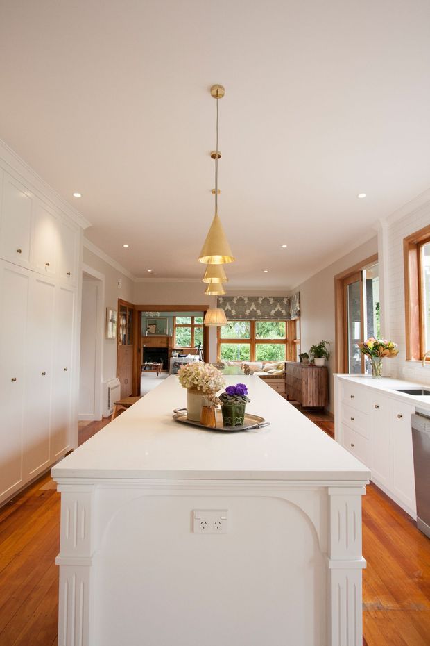 Bright, refined kitchen centred on a large white island with decorative paneled ends and a pale benchtop. The room features extensive white built-in cabinetry, timber flooring, warm timber-framed windows, recessed ceiling lights, and a row of brass/gold pendant lights. A patterned window treatment and floral arrangements add decorative detail, while an adjoining living or dining area is visible beyond the kitchen.