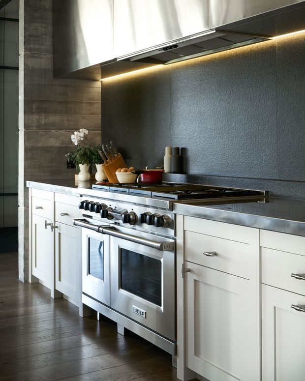A close-up view of a refined kitchen cooking zone featuring a large stainless-steel range cooker, white shaker-style cabinetry, dark textured backsplash, dark wood flooring, stainless-steel upper cabinetry, and warm linear under-cabinet lighting. Minimal countertop accessories and a vase of orchids add restrained decorative detail.