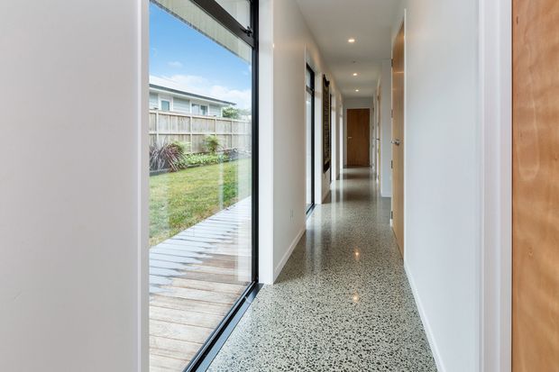 A bright, narrow residential hallway with polished speckled terrazzo flooring, white walls, recessed ceiling lights, and timber doors. Full-height glazing on the left opens views to a small lawn, timber decking, and a fenced neighbouring property, while the corridor leads toward a closed timber door at the far end.