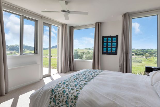 Bright contemporary bedroom with expansive windows overlooking a green rural landscape. The room features a white upholstered bed with white bedding and a blue-green floral runner, pale curtains, light carpet, white walls, recessed ceiling lights, and a white ceiling fan. A colourful framed artwork provides a small accent on the wall.