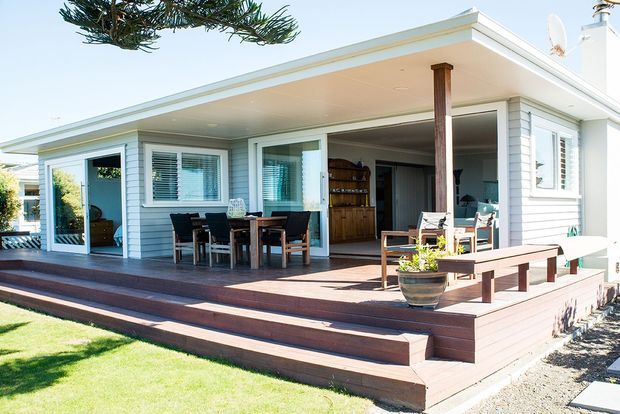 A bright single-storey coastal-style home viewed from the garden, featuring a broad covered deck with timber decking, multiple steps, outdoor dining furniture, bench seating, white weatherboard cladding, wide glazed openings, and a low-pitched flat roof. Interior living and dining areas are visible through the open doors.