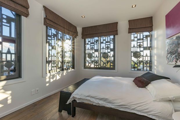 Bright contemporary bedroom with a large bed positioned in a corner, white bedding, dark wood furnishings, and multiple tall windows fitted with patterned metal security screens and brown Roman blinds. Warm timber flooring and recessed ceiling lights create a clean, refined interior, with strong geometric window shadows adding visual interest.