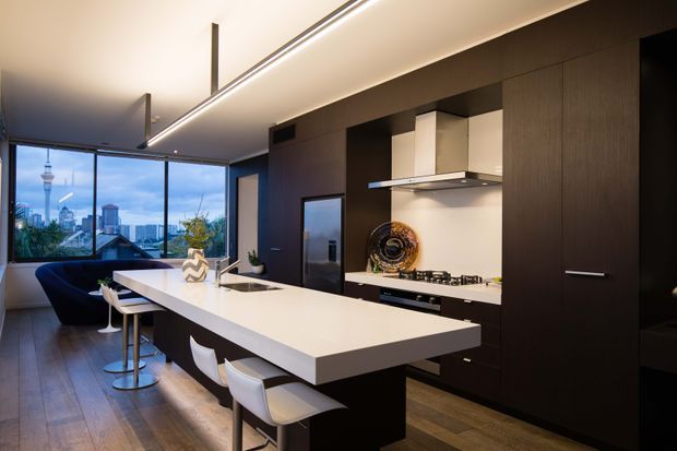 Contemporary open-plan kitchen with a large white island and integrated seating, dark timber-toned full-height cabinetry, stainless steel appliances, and a linear ceiling light. Floor-to-ceiling glazing opens the view toward an urban skyline, while a dark lounge area and dining stools extend beyond the kitchen.