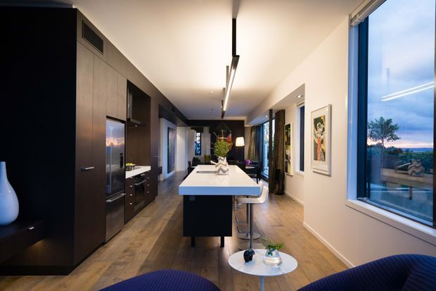 A long, contemporary open-plan kitchen and living area viewed along a central island. Dark timber-toned full-height cabinetry lines the left wall, with integrated stainless-steel appliances and a pale benchtop. The island has a white surface, dark base, and two bar stools. Warm timber flooring, white walls and ceiling, linear pendant lighting, framed artwork, and deep blue lounge seating create a refined interior. Large windows on the right provide expansive outdoor views.