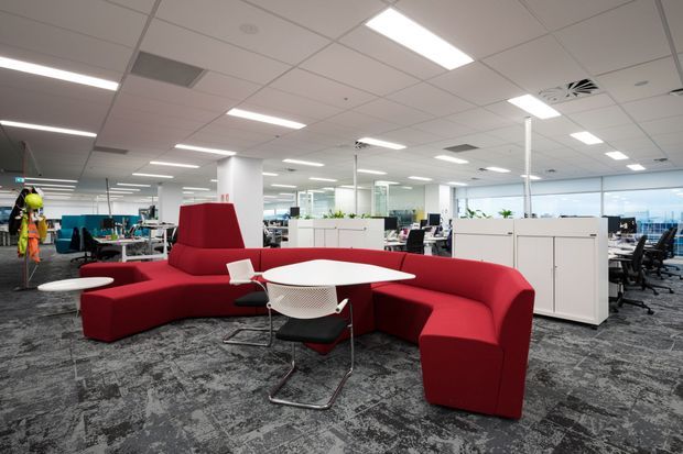 An expansive contemporary open-plan office with low white acoustic tile ceilings, extensive workstations, glass partitions, white storage units, and patterned grey carpet. A bold red modular lounge seating arrangement surrounds a white round table in the foreground, with task chairs and desks extending across the bright commercial workspace.