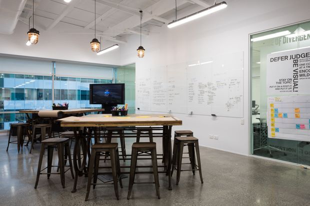 A bright contemporary collaborative office or studio space with long communal timber worktables, stools, a large wall-mounted whiteboard, extensive internal glazing, and exposed services in the ceiling. Pendant lights and linear suspended fixtures illuminate the polished concrete floor, while a glass-partitioned meeting or work area is visible at right.