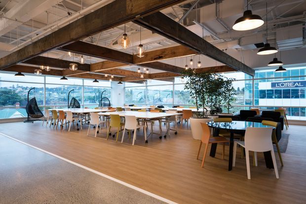 A spacious contemporary commercial office breakout and dining area with extensive floor-to-ceiling glazing, open-plan communal tables, lounge seating, and a mix of timber and colourful chairs. Exposed dark timber beams and exposed services span the ceiling, complemented by suspended pendant lights. The space overlooks an urban setting and includes indoor planting, with a visible L'Oréal building outside.