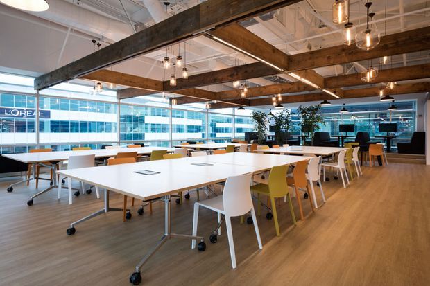 Open-plan commercial office or collaborative workspace with large floor-to-ceiling windows, rows of movable white tables and colourful task chairs, timber-look flooring, exposed white ceiling services, and substantial dark timber beams with pendant lighting. The space has a bright, contemporary-industrial character and overlooks an urban commercial setting.