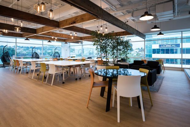 A bright, open-plan commercial office breakout and collaboration area with extensive glazing and views to neighboring buildings. The space features warm timber-look flooring, exposed services and structural ceiling beams, numerous shared dining and work tables, colorful mixed chairs, black pendant lighting, lounge seating, and indoor greenery. The overall design is contemporary with industrial-inspired exposed structure and a relaxed workplace hospitality feel.