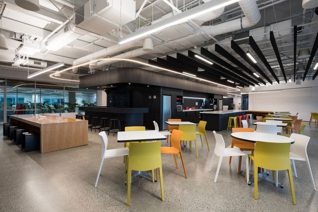 Open-plan commercial office hospitality and breakout area with a café-style black service counter, communal timber island, numerous colourful tables and chairs, polished exposed services, linear pendant lighting, and a terrazzo-like floor. The space has an industrial contemporary fit-out with visible ductwork and structural ceiling services.