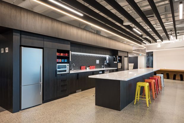 A spacious contemporary commercial-style kitchen with dark timber-look cabinetry, stainless-steel appliances, a long pale stone island bench, open shelving, and colourful bar stools. The space features polished concrete or terrazzo-style flooring, linear pendant lighting, exposed black ceiling beams, and visible ductwork, creating an industrial yet refined workplace fit-out.