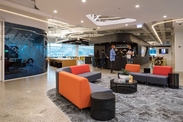 A contemporary commercial office reception and lounge area with colourful upholstered sofas, a low round coffee table, patterned carpet, polished concrete flooring, and a black built-in bar or breakout hub. The space features exposed services, suspended linear and sculptural lighting, curved glazed meeting-room walls, and an open-plan workspace beyond.