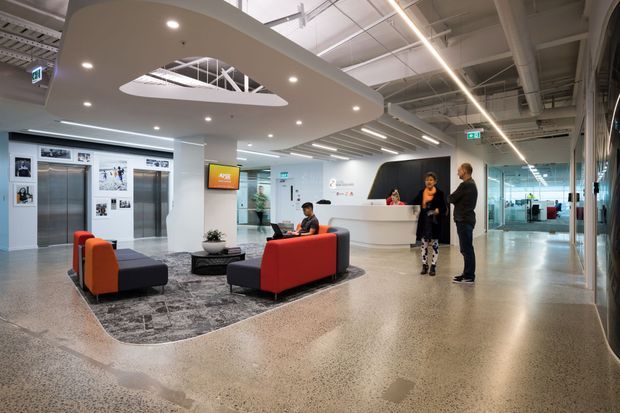 A contemporary commercial reception and waiting area with a white curved reception desk, lounge seating, polished speckled concrete or terrazzo flooring, glass partitions, elevator doors, and an exposed industrial-style ceiling with linear lighting. A large suspended curved ceiling feature with an oval opening creates a focal point above the seating area.
