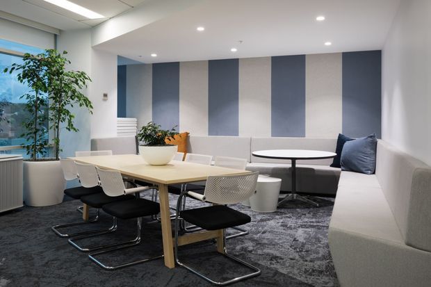 A bright contemporary office breakout and meeting area with a large light-wood conference table, white cantilever chairs, a round table with booth seating, and a long upholstered sofa. The space features a dark patterned carpet, blue and pale neutral vertical wall panels, recessed ceiling lights, indoor planting, and glazing along one side.