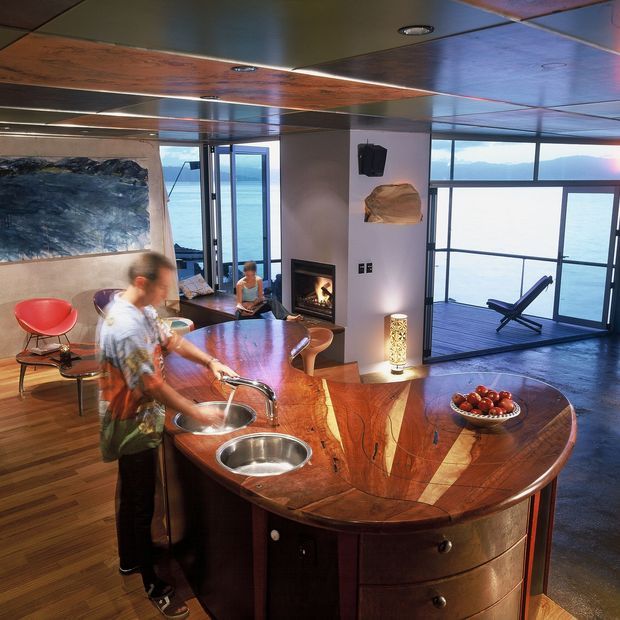 Open-plan kitchen and living space with a sculptural curved timber island featuring twin sinks and integrated cabinetry. The room has warm timber flooring, a reflective paneled ceiling, a fireplace, contemporary furniture, and expansive glazing overlooking a body of water with an outdoor deck and lounge chair.