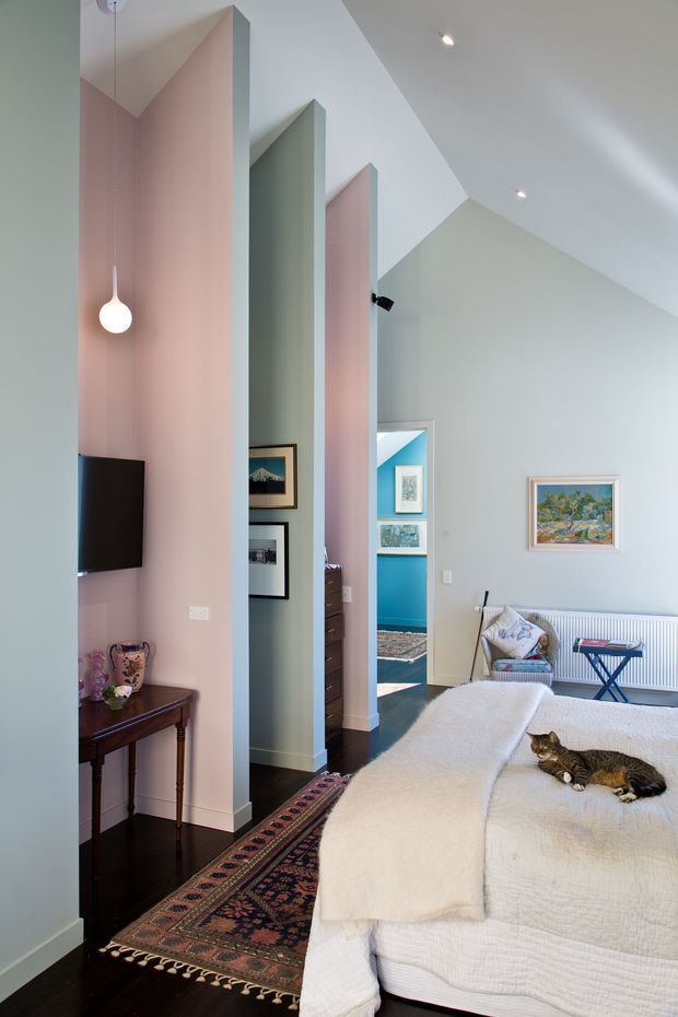 A light-filled bedroom with a high, angular ceiling and layered pastel partition walls in blush pink, sage green, and pale blue. The room contains a large upholstered bed with a white textured cover, a small blue folding side table, a radiator, framed artwork, a dark timber console, wall-mounted television, patterned runner rug, and a pendant light. Openings between the partitions reveal adjoining circulation and rooms, including a bright blue accent wall.