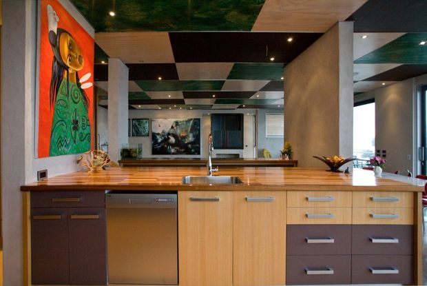 Contemporary open-plan kitchen dominated by a large central island with a timber benchtop, stainless-steel sink and integrated dishwasher. The island combines light timber cabinetry with dark brown drawer fronts and a dark-brown cabinet section. Behind it is a second timber counter, with concrete-look structural columns, a highly distinctive checkerboard ceiling of dark, light timber and green panels, recessed lighting, and colourful artwork. Dining and living areas are visible beyond the kitchen.