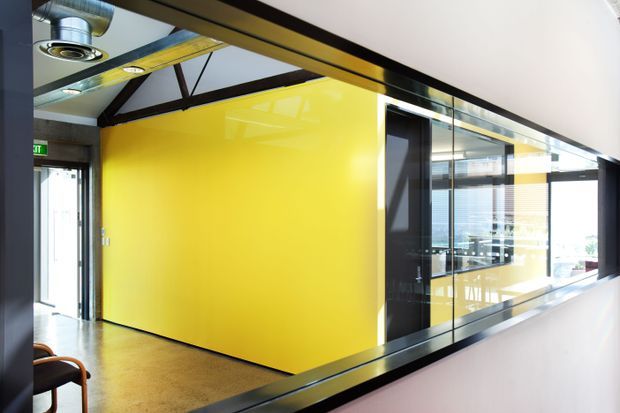 A contemporary commercial interior viewed along a glass-partitioned corridor. A vivid yellow enclosed meeting or office volume is framed by black metal and extensive glazing, with exposed dark roof trusses, polished metal ductwork, recessed circular ceiling lights, and a polished concrete or terrazzo floor. Reflections reveal additional workspaces beyond the glass. The space has a bold, high-contrast industrial-office character.