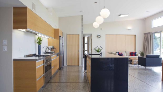 Bright open-plan interior combining a modern kitchen and living room. The kitchen features warm timber cabinetry, a dark benchtop, stainless-steel appliances and a polished concrete or terrazzo-like floor. A black kitchen island divides the kitchen from the living area, which includes neutral seating, timber wall panelling, indoor plants and spherical pendant lights. High-level windows and a light neutral palette create an airy, contemporary atmosphere.