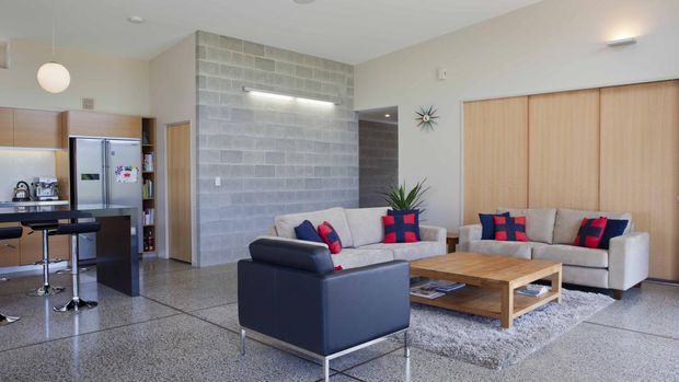 Open-plan living room with a compact kitchen at left, polished concrete block feature wall, pale timber sliding doors, and mid-century-inspired sofas and armchairs arranged around a timber coffee table. The room has polished concrete flooring, neutral walls, abundant natural light, and colourful geometric cushions.