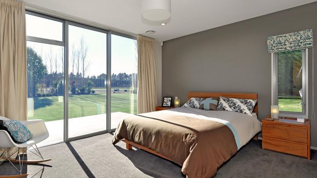 Bright contemporary bedroom with a low timber bed, neutral brown bedding, grey carpet, and a dark grey feature wall. Floor-to-ceiling glazing and a large window provide views of landscaped grounds, while a patterned Roman blind and light curtains frame the windows. A timber bedside table, simple pendant light, and accent chair complete the room.