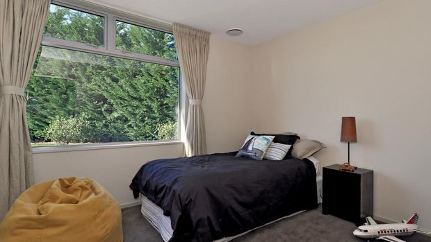 A compact, simply furnished bedroom with a double bed dressed in dark navy bedding, neutral upholstered curtains, a black bedside table, and a warm brown table lamp. Large windows provide views of dense greenery, while a mustard-yellow beanbag and a small toy airplane add casual character.