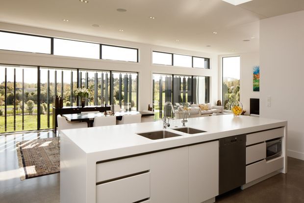 Bright, spacious contemporary open-plan kitchen with a large white island, integrated double sinks, dishwasher and oven, opening onto a dining and living area. Floor-to-ceiling black-framed glazing provides expansive views of the landscaped outdoors, while recessed ceiling lighting and white cabinetry create a clean, refined interior.