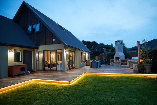 Dusk exterior view of a contemporary gabled house opening onto a broad timber deck and lawn. The dark vertical-clad upper volume contrasts with pale rendered walls and extensive glazed doors. Integrated warm LED strip lighting outlines the deck edge, while a substantial outdoor fireplace and seating area occupy the far side of the terrace.