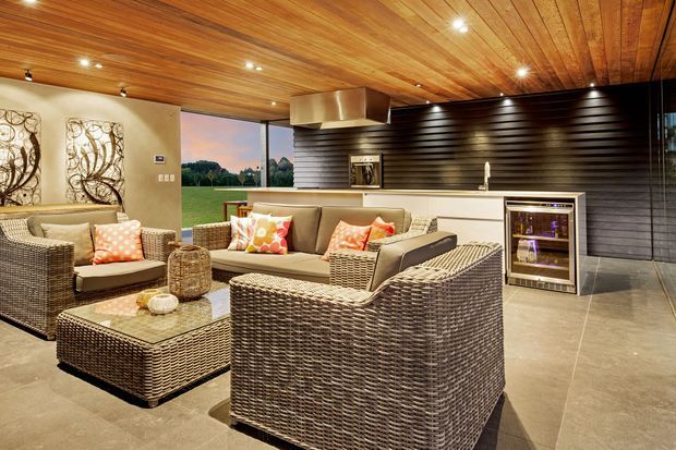 A refined open-plan entertaining space combining a lounge area and compact kitchen/bar. The room features extensive warm timber ceiling lining, dark horizontal wall cladding, light cabinetry, a stone-look island benchtop, polished concrete or large-format grey flooring, woven outdoor-style seating, and a built-in wine fridge. Full-height glazing opens views toward a green landscape.