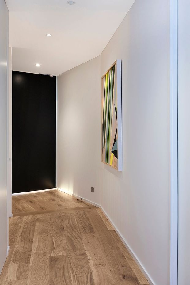 A minimalist interior hallway or entry passage with pale painted walls, recessed ceiling lights, and warm natural timber flooring. A tall black door or dark wall panel anchors the far end, while a colourful abstract artwork provides the main decorative feature on the side wall. Bright glazing or a white-framed opening is visible along the right edge.