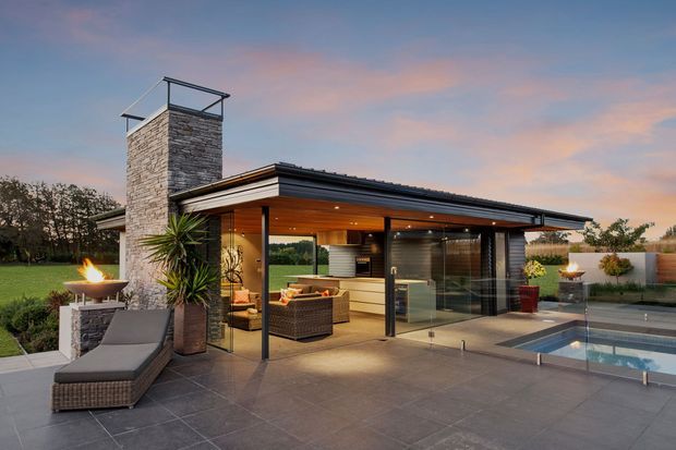 A modern single-storey pavilion-style home opening onto a paved terrace and swimming pool at dusk. The building features a broad low-pitched roof with deep eaves, extensive floor-to-ceiling glazing, warm timber soffits, dark metal framing, and a prominent stacked-stone chimney. Outdoor lounge furniture, fire bowls, landscaped planting, and the pool create a refined indoor-outdoor entertaining area.