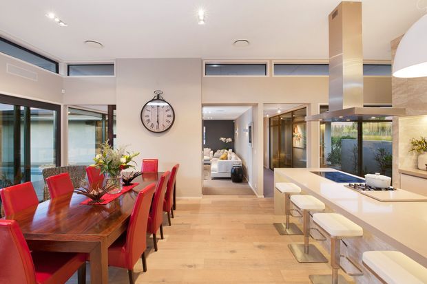 A bright, open-plan contemporary interior combining a dining area and kitchen. The dining space features a substantial dark timber table with red upholstered chairs, while the kitchen has a white island with integrated appliances, a raised pale stone or solid-surface benchtop, and white stools. Full-height glazing and high-level windows bring in abundant light, with a hallway and living area visible beyond. A large stainless-steel rangehood provides a strong vertical focal point.