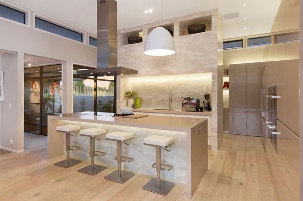 Contemporary open-plan kitchen with a large stone-clad island, four white upholstered bar stools, integrated cooktop and rangehood, illuminated splashback shelving, full-height cabinetry, and a stainless-steel refrigerator. Light timber flooring, neutral finishes, skylight-like clerestory windows, and pendant lighting create a bright, refined space.