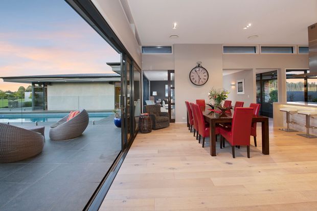Open-plan contemporary interior with a dining area and kitchen extending toward a living space, framed by expansive floor-to-ceiling glazing and opening onto a covered terrace with a swimming pool. The room features pale timber flooring, white walls and ceiling, a dark-framed glazed wall, a large timber dining table with vivid red upholstered chairs, and a light-toned kitchen island. The outdoor area is visible through the fully glazed opening.
