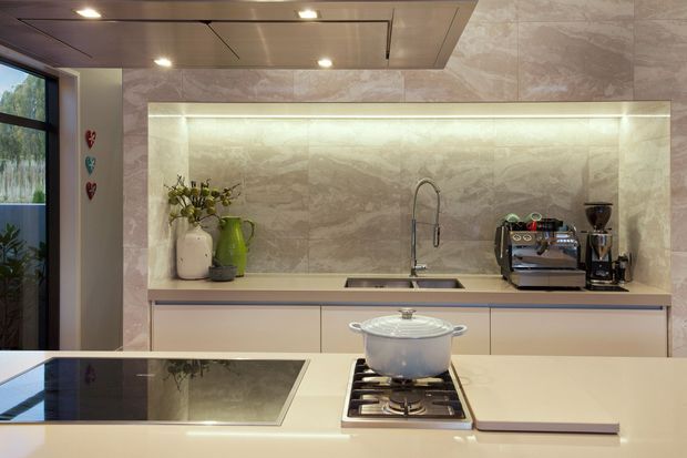 Contemporary kitchen with a pale stone-look full-height splashback and illuminated recessed display niche, streamlined light cabinetry, integrated sink, induction cooktop and a gas burner on the island. A coffee station and decorative vessels sit along the rear benchtop, with dark-framed glazing at the side.