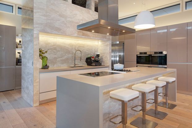 Contemporary open-plan kitchen with a large waterfall-edge island, integrated appliances, pale glossy cabinetry, stone-look splashback and benchtops, and four upholstered bar stools. A stainless-steel rangehood forms a central feature above the island cooktop, while concealed under-cabinet lighting and pendant lighting create a polished, warm interior.