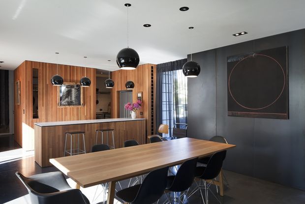 A contemporary open-plan dining and kitchen interior with a long timber dining table, dark upholstered dining chairs, a timber-lined kitchen and island, black pendant lights, and a dark feature wall with artwork. Floor-to-ceiling glazing and sheer curtains bring in natural light.
